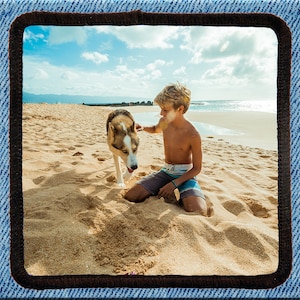 May include: A young boy sits on a sandy beach with a brown and white dog. The boy is wearing blue swimming trunks and has light brown, wavy hair. The dog is looking at the boy and has its tongue sticking out. The beach is empty and the sea is in the background.