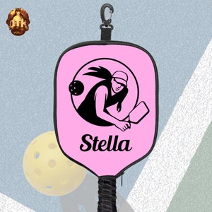 Puede incluir: Raqueta de pickleball rosa con una silueta negra de un jugador sosteniendo una raqueta y una pelota. La raqueta tiene una correa negra y una cremallera negra. El nombre "Stella" est&aacute; impreso en la raqueta.