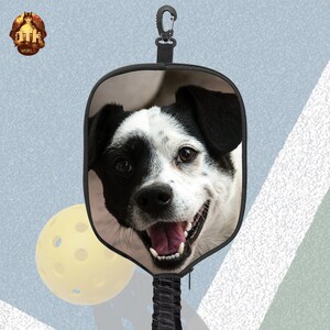 Puede incluir: Una raqueta de pickleball negra y beige con la imagen de un perro blanco y negro. La raqueta tiene un mango negro con un mosquet&oacute;n negro.