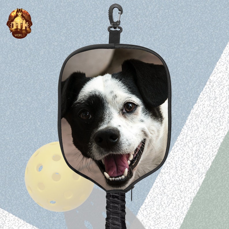 Puede incluir: Una raqueta de pickleball negra y beige con la imagen de un perro blanco y negro. La raqueta tiene un mango negro con un mosquet&oacute;n negro.