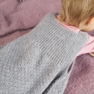 Può includere: Un gilet grigio lavorato a maglia con un motivo strutturato. L'indumento senza maniche è mostrato su un bambino, con una camicia a righe rosa e bianca visibile sotto. Il gilet è realizzato in un materiale morbido, possibilmente lana.