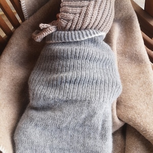 May include: A gray knitted baby sleeping bag with a brown knitted hat. The baby is swaddled in the sleeping bag and is lying on a brown blanket.
