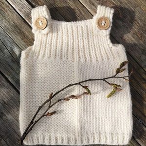 Könnte beinhalten: Ein weißes, ärmelloses Strickoberteil mit Holzknöpfen. Das Oberteil liegt auf einer Holzoberfläche mit einem kleinen Zweig mit Blättern.