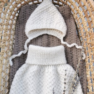 Può includere: Un completo per neonato lavorato a maglia, composto da un cappellino e un maglione con collo alto, di colore crema chiaro. Il completo è esposto in un cestino di vimini, con una coperta marrone sottostante. Un rametto di lavanda aggiunge un tocco di colore.