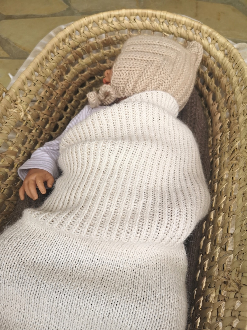 May include: A white knitted baby sleeping bag with a matching knitted hat. The baby is lying in a woven basket.