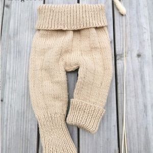 Può includere: Un paio di pantaloni da bambino beige a maglia con fascia in vita ripiegata e polsini a coste. I pantaloni sono realizzati in un filato morbido e strutturato e sono esposti su una superficie di legno. Il testo "German & English" è nell'angolo in alto a sinistra.