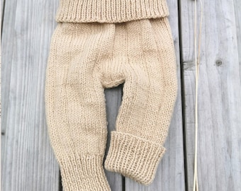 May include: Beige knitted baby trousers with a folded waistband and ribbed cuffs. The soft, textured yarn trousers are displayed on a wooden surface. The text "German & English" is in the upper left corner. The trousers are approximately 40 cm long.