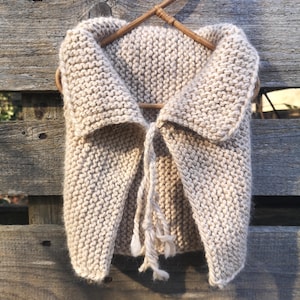 Peut inclure: Un gilet en tricot beige avec un col châle et une fermeture à nouer. Le gilet est suspendu à un cintre en bois.