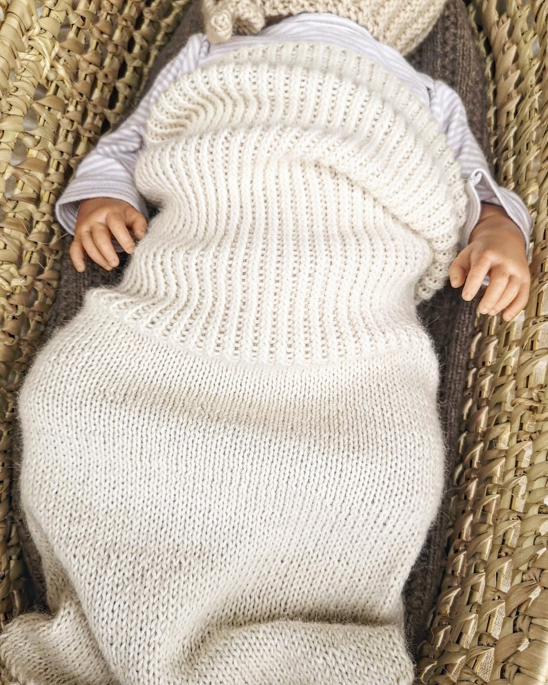 May include: A white knitted baby sleeping bag with a ribbed texture. The sleeping bag is folded over the baby's chest and is lying in a woven basket.