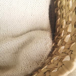 May include: A close-up of a woven wicker basket with a cream-colored knitted blanket draped over the edge.