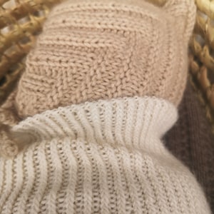 May include: A close-up of a beige knitted hat with a white knitted turtleneck sweater underneath. The hat has a fold-over brim and ear flaps.