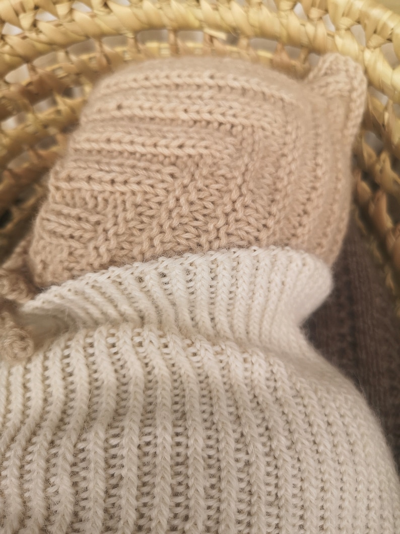 May include: A close-up of a beige knitted hat with a white knitted turtleneck sweater underneath. The hat has a fold-over brim and ear flaps.