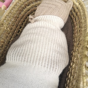 May include: A white knitted baby doll sleeping in a woven basket. The doll is wearing a beige knitted hat.