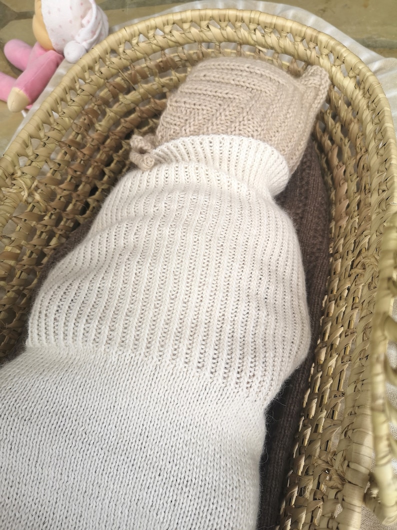 May include: A white knitted baby doll sleeping in a woven basket. The doll is wearing a beige knitted hat.