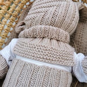 May include: A beige knitted set consisting of a hat with small ears, a textured cowl or collar piece, and a ribbed garment, all worn over a white long-sleeved shirt. The knitwear features a consistent light tan color and varied stitch patterns.