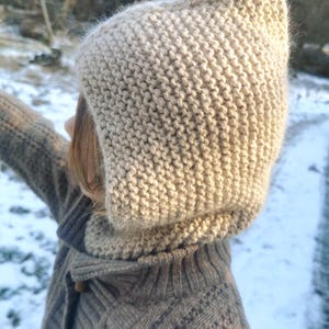 Peut inclure: Une capuche tricotée beige avec un sommet pointu, fabriquée à partir d'un fil texturé avec une finition duveteuse visible. La capuche présente un motif au point mousse et couvre la tête et le cou, offrant une forme structurée et pointue.