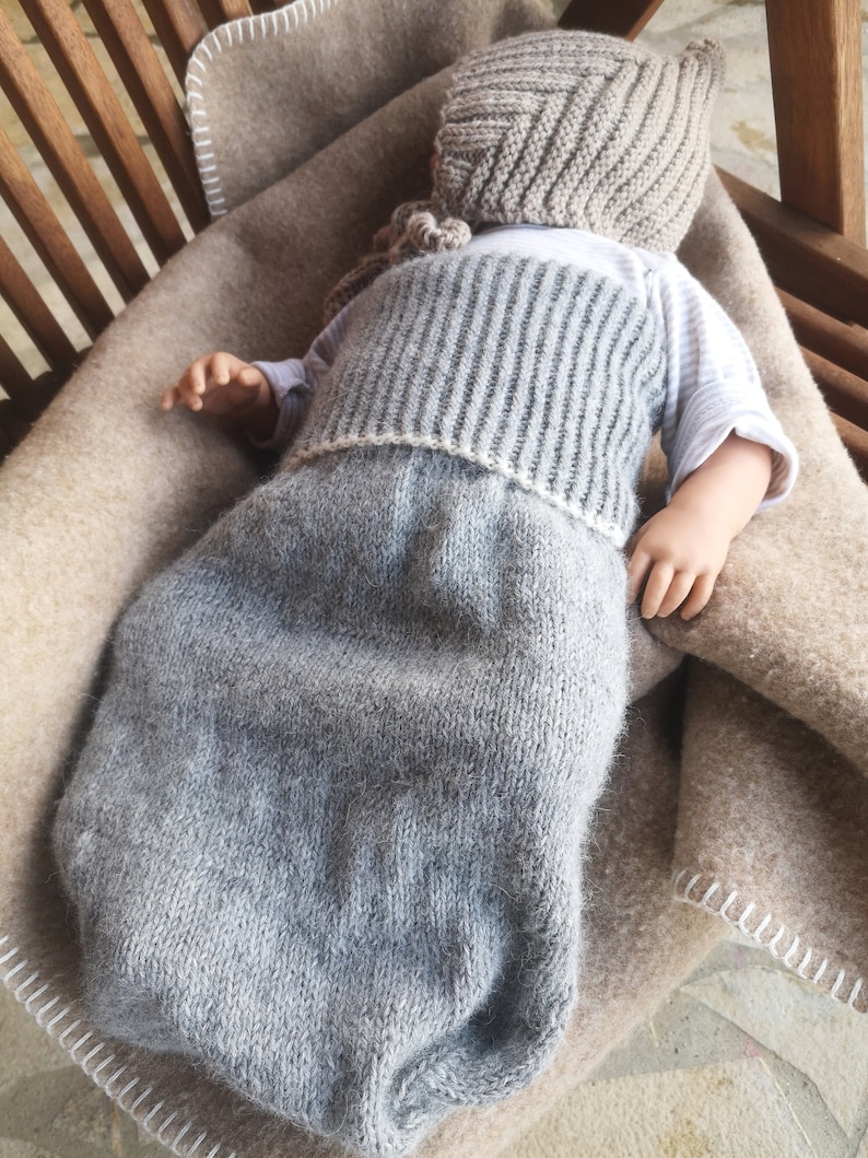 May include: A gray knitted baby sleeping bag with a matching gray knitted hat. The sleeping bag is lying on a brown blanket.