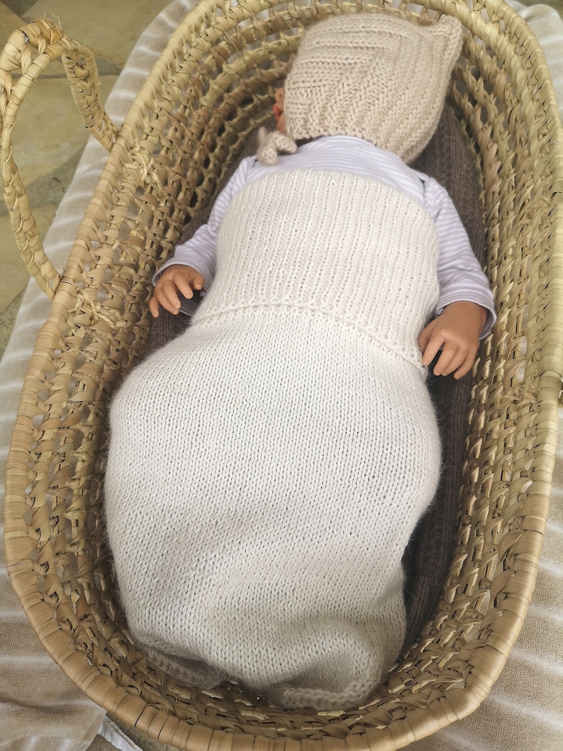 May include: A cream-colored knitted baby sleeping bag with a matching knitted hat. The sleeping bag is in a woven basket.