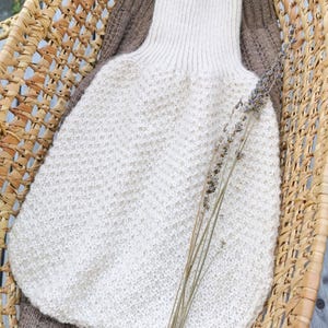 Könnte beinhalten: Ein cremefarbener, handgestrickter Pullover mit hohem Kragen und strukturiertem Strickmuster, in einem Weidenkorb. Ein brauner Pullover liegt darunter. Getrocknete Lavendelzweige liegen obenauf.