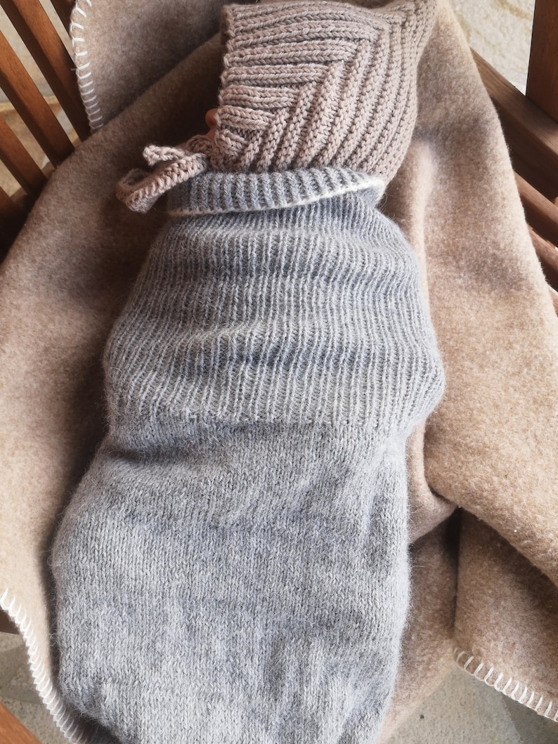 May include: A gray knitted baby sleeping bag with a brown knitted hat. The baby is swaddled in the sleeping bag and is lying on a brown blanket.
