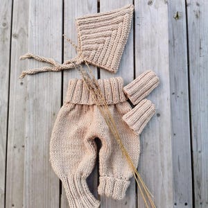 May include: A beige knitted set consisting of a pointed bonnet with braided ties, high-waisted trousers with a wide ribbed waistband and cuffs, and two matching ribbed wrist cuffs. The items feature a consistent chunky knit texture throughout.