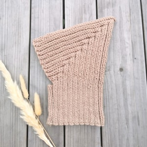 Op de afbeelding: Een lichtbruine gebreide bivakmuts, liggend op een houten oppervlak. De bivakmuts is gemaakt van een zacht, getextureerd garen en heeft een ribgebreid ontwerp. De hoofdbedekking is ontworpen om het hoofd en de nek te bedekken.