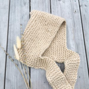 Puede incluir: Una bufanda beige tejida descansa sobre una superficie de madera envejecida, con acentos florales secos. La bufanda está hecha de un hilo grueso y texturizado, y está doblada para mostrar su longitud. El color neutro y el diseño sencillo sugieren un accesorio acogedor y hecho a mano.