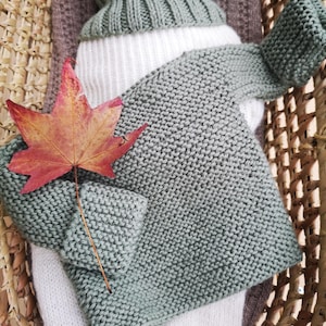 Könnte beinhalten: Ein salbeigrüner gestrickter Baby-Pullover mit langen Ärmeln und einem gerippten Kragen. Der Pullover liegt in einem geflochtenen Korb mit einer braunen und weißen Decke und einem roten Ahornblatt.