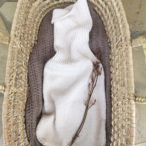 May include: A white knitted turtleneck sweater with a brown knitted sweater underneath, both folded and placed in a woven basket.
