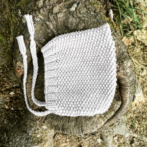 Könnte beinhalten: Eine weiße gestrickte Babyhaube mit einem strukturierten Stichmuster und langen Bändern.