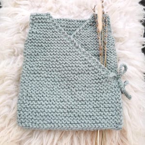 May include: A pale blue knitted wrap top with a cross-over design and tie closure. The top is made of a soft, textured yarn and is displayed on a white, fluffy surface. Dried flowers are tucked into the wrap.