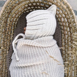 Könnte beinhalten: Eine weiße gestrickte Baby-Mütze mit Ohrenklappen, eingewickelt in eine passende weiße gestrickte Decke, die in einem geflochtenen Korb liegt.