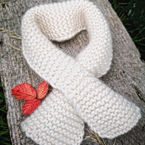 Könnte beinhalten: Ein cremefarbener Strickschal mit kraus rechts gestricktem Muster und einer weichen, flauschigen Textur. Der Schal ist in einer Schlaufenform angeordnet und wird von drei kleinen, leuchtend roten Blättern am unteren linken Rand begleitet.