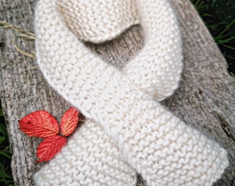 May include: A cream-colored knitted scarf featuring a garter stitch pattern with a soft, fuzzy texture. The scarf is arranged in a loop shape and is accompanied by three small, vibrant red leaves positioned near the bottom left edge.