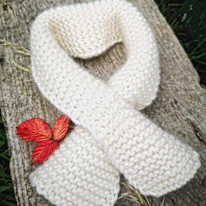 Puede incluir: Una bufanda de punto blanco con una textura suave y peluda. La bufanda está doblada en forma de bucle y está sobre una superficie de madera.