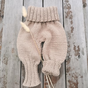 Può includere: Un paio di pantaloni da neonato beige lavorati a maglia con una vita alta e ripiegata e polsini a coste. I pantaloni sono realizzati in un filato morbido e strutturato e sono esposti su una superficie di legno invecchiato. Un rametto di fiori secchi aggiunge un tocco naturale.