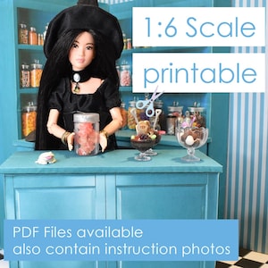 Puede incluir: Una muñeca con un vestido negro y un sombrero de bruja se encuentra frente a un mostrador de tienda azul en miniatura. El mostrador tiene frascos de caramelos y un sundae de helado en miniatura. El texto "1:6 Scale printable" se muestra encima del mostrador. El texto "PDF Files available also contain instruction photos" se muestra debajo del mostrador.