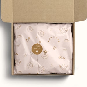 May include: A cardboard box with a pink and white patterned tissue paper inside. The tissue paper has a gold design of moons, stars, mountains, and hands. A gold sticker on the tissue paper reads "MADE WITH" and has a heart symbol.