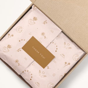 May include: A brown cardboard box with a pink and gold patterned tissue paper inside. The tissue paper has a repeating pattern of flowers, moons, stars, and hands. A small gold card with the text "THANK YOU!" is placed on top of the tissue paper.