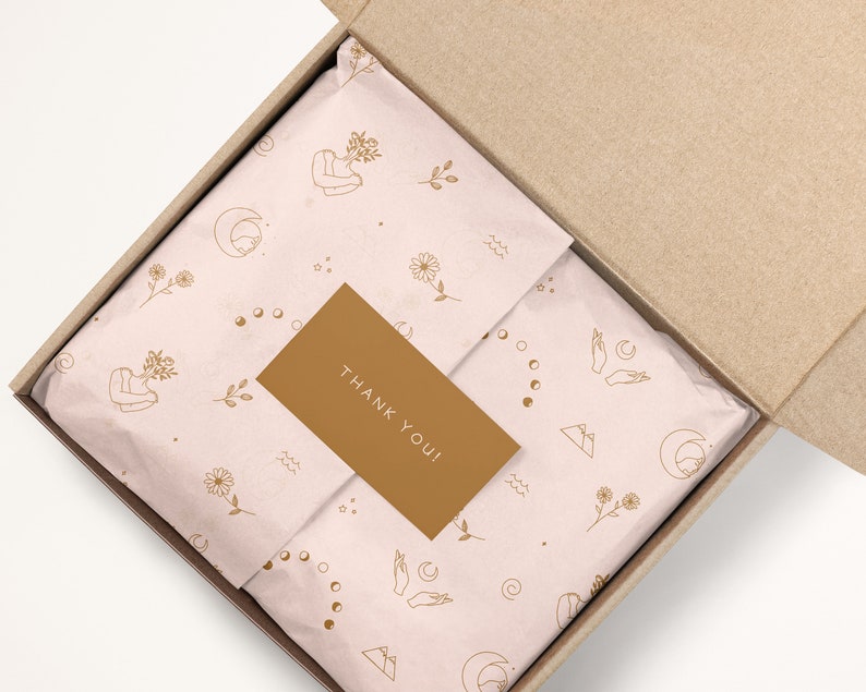 May include: A brown cardboard box with a pink and gold patterned tissue paper inside. The tissue paper has a repeating pattern of flowers, moons, stars, and hands. A small gold card with the text "THANK YOU!" is placed on top of the tissue paper.