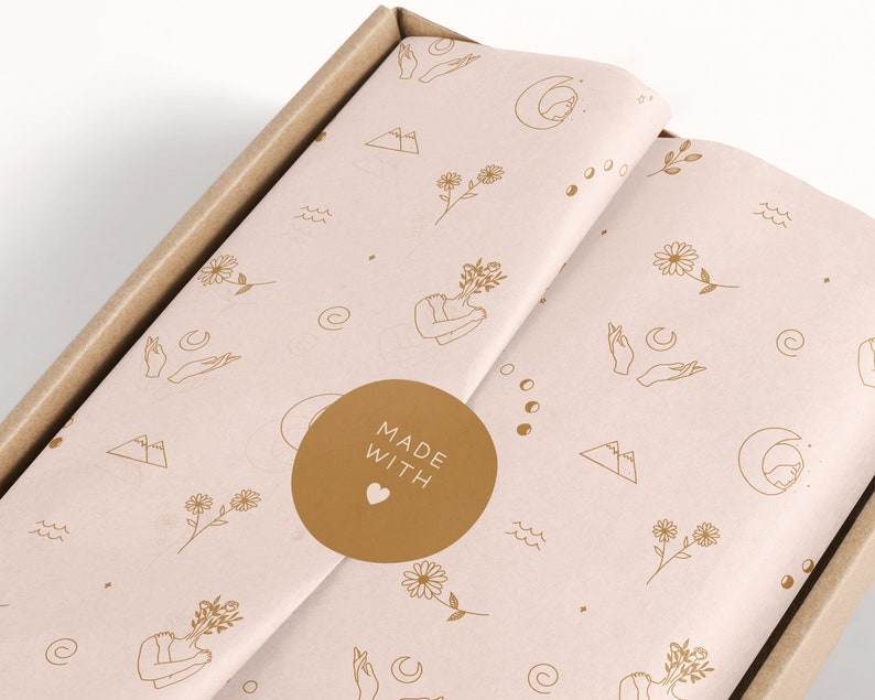 May include: A close-up of a brown cardboard box with a pink and gold patterned tissue paper inside. The tissue paper has a repeating pattern of flowers, moons, hands, and mountains. A gold circle sticker with the text "MADE WITH HEART" is on the tissue paper.