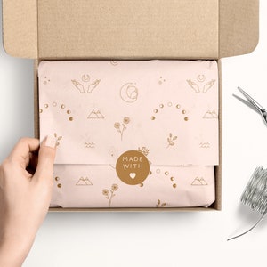 May include: A light pink gift box with a brown cardboard lid is open, revealing tissue paper with a delicate pattern of moons, stars, mountains, and flowers. The tissue paper has a round sticker that says "MADE WITH" and a heart symbol.