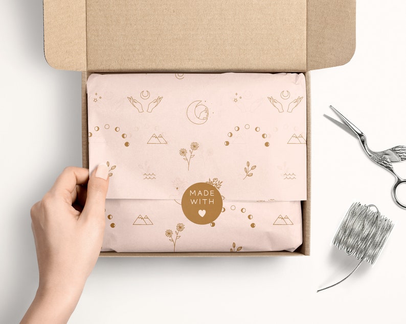May include: A light pink gift box with a brown cardboard lid is open, revealing tissue paper with a delicate pattern of moons, stars, mountains, and flowers. The tissue paper has a round sticker that says "MADE WITH" and a heart symbol.