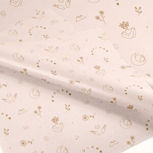 May include: A pale pink wrapping paper with a repeating pattern of gold line drawings. The pattern includes moons, stars, mountains, flowers, and hands in a prayer position.