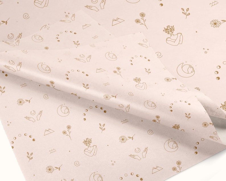May include: A pale pink wrapping paper with a repeating pattern of gold line drawings. The pattern includes moons, stars, mountains, flowers, and hands in a prayer position.