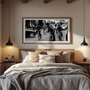 Puede incluir: Una fotografía en blanco y negro con marco rectangular que muestra una concurrida escena callejera con varias personas montando bicicletas entre coches. La obra está montada en una pared clara sobre una cama con almohadas y una manta.