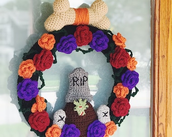 May include: A crocheted wreath with a bone-shaped bow, red, orange, purple, and green flowers, and a small gray tombstone with the word "RIP" on it.