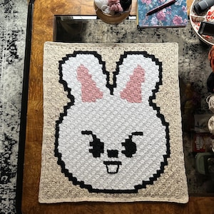 May include: A crocheted square with a cartoon bunny design. The bunny has white fur, pink ears, and black facial features. The square is framed by a beige border.