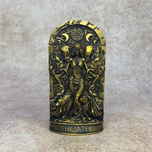 May include: A gold-colored statue of the Greek goddess Hecate, depicted with three heads and holding torches. The statue is standing on a base with the word "Hecate" inscribed on it.