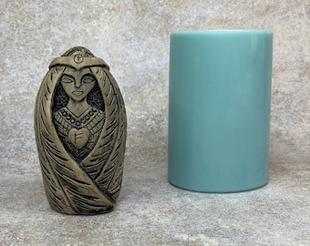 May include: A small, detailed, gold-colored ceramic sculpture of a woman wearing a crown and a flowing robe. The sculpture is standing on a light gray surface next to a teal-colored candle.